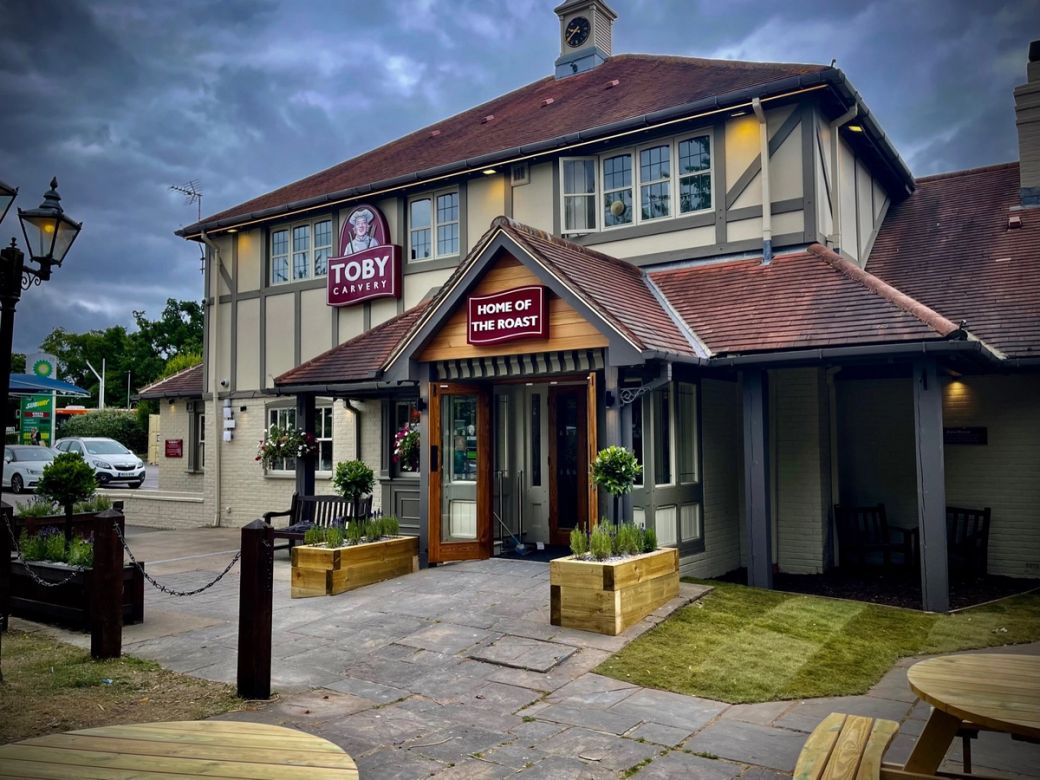 The image shows a traditional pub building with a prominent entrance and decorative outdoor seating. The structure features a gabled roof and is surrounded by greenery, creating a welcoming atmosphere.