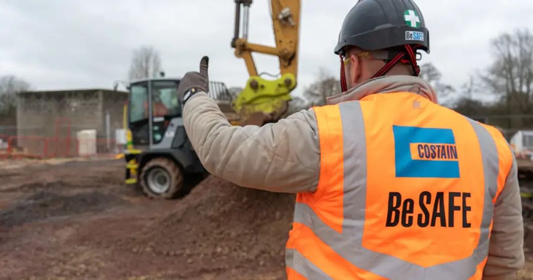 A construction worker in a high-visibility vest gives a thumbs up while standing on a construction site. The background features heavy machinery and earth-moving equipment.