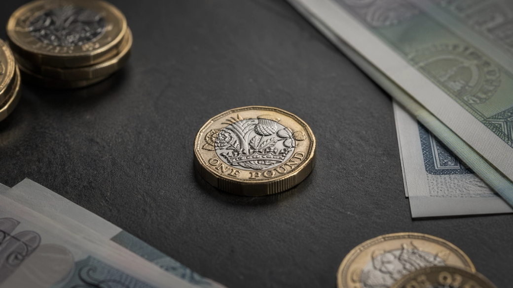The image features a close-up view of a one-pound coin placed on a dark surface, surrounded by various denominations of paper currency and additional coins. The composition highlights the details of the coin alongside the notes, emphasizing currency.