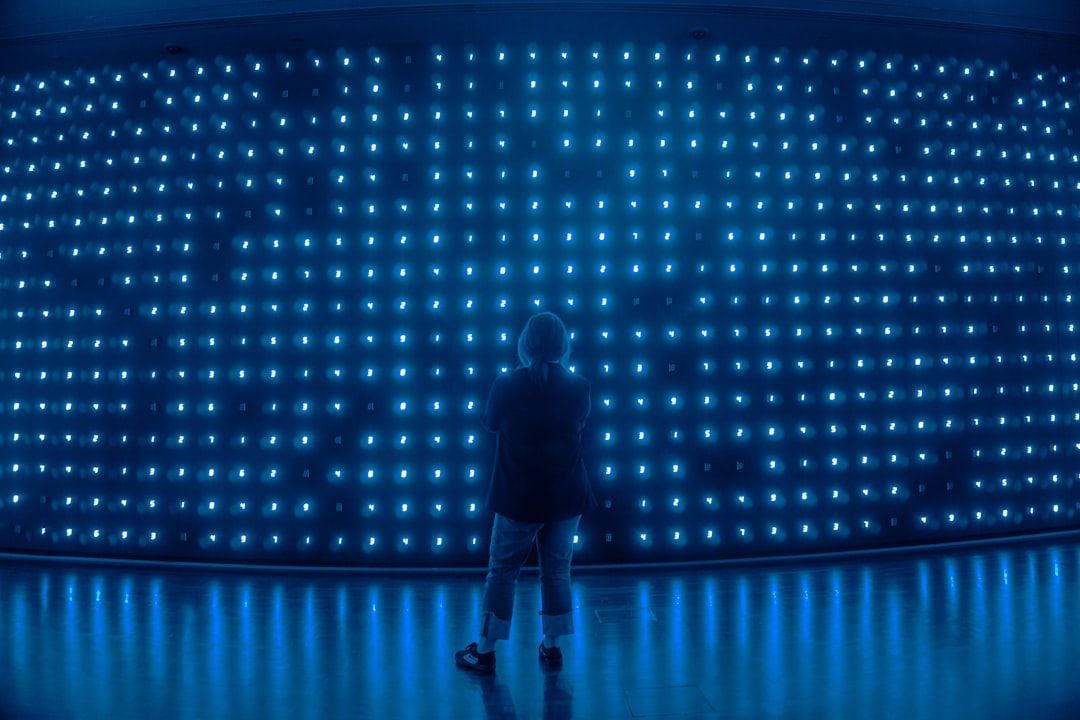 The image depicts a person standing in front of a large wall of glowing blue lights. The ambient illumination creates a striking visual effect, emphasizing the contrast between the subject and the background.
