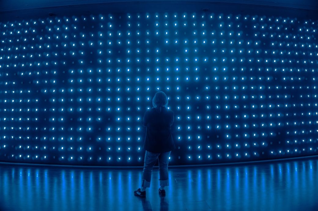 The image depicts a person standing in front of a large wall of glowing blue lights. The ambient illumination creates a striking visual effect, emphasizing the contrast between the subject and the background. — Credit: Photo by Y K on Unsplash