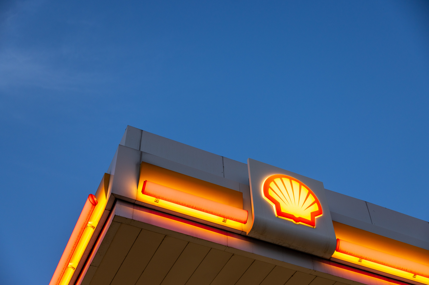 The image features the illuminated signage of a Shell gas station against a clear blue sky. The iconic Shell logo is prominently displayed at the top, highlighting the brand's presence in the energy sector.