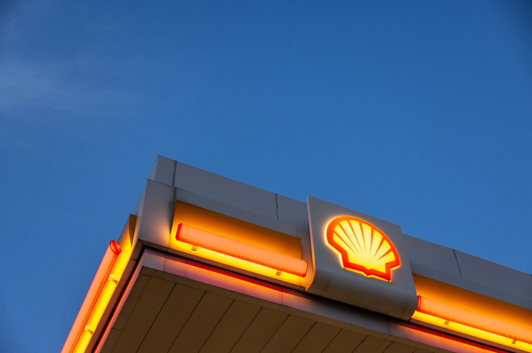 The image features the illuminated signage of a Shell gas station against a clear blue sky. The iconic Shell logo is prominently displayed at the top, highlighting the brand's presence in the energy sector.