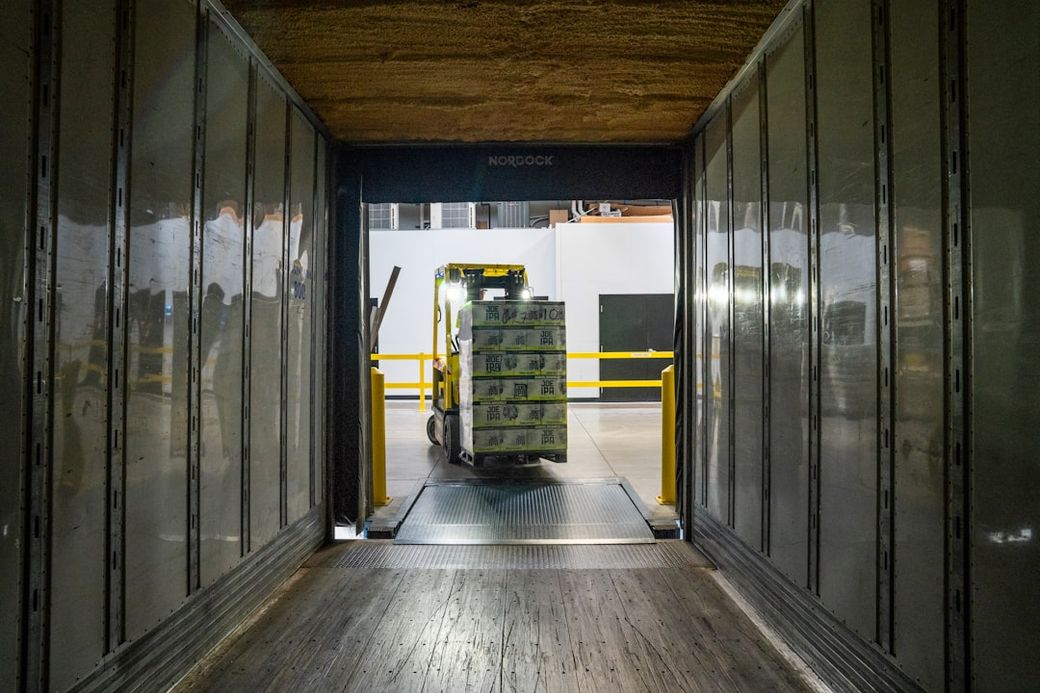 A forklift is seen transporting a stack of boxes out of a trailer loading dock. The interior of the dock features reflective walls and industrial design elements. — Credit: Photo by Elevate on Unsplash