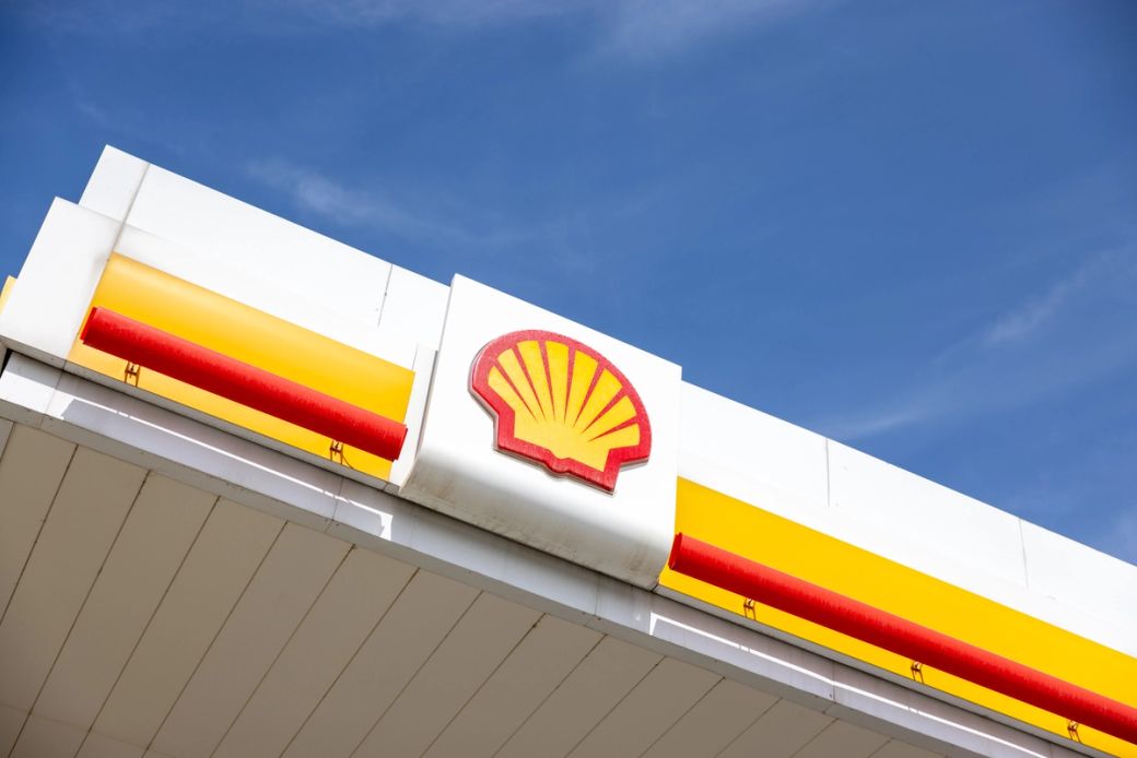 The image depicts the exterior signage of a Shell gas station against a clear blue sky. The logo, featuring the iconic shell design in red and yellow, is prominently displayed.