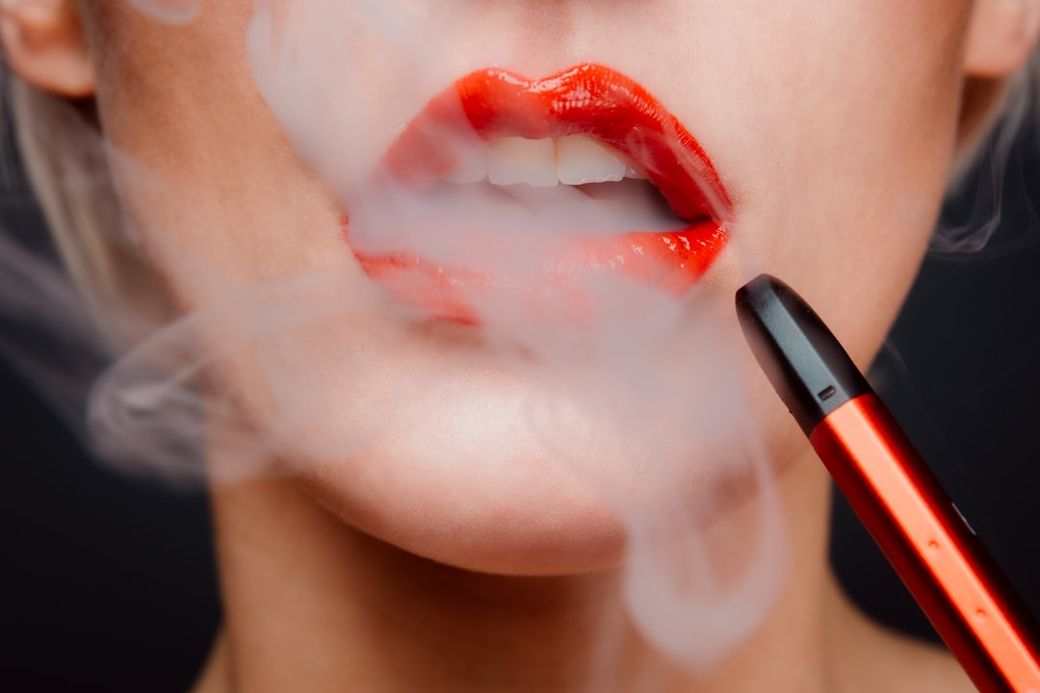 A close-up image of a woman's lips adorned with bright red lipstick, exhaling vapor from a vape pen. The background is dark, emphasizing the vivid colors and the smoke effect. — Credit: Photo by Chiara Summer on Unsplash