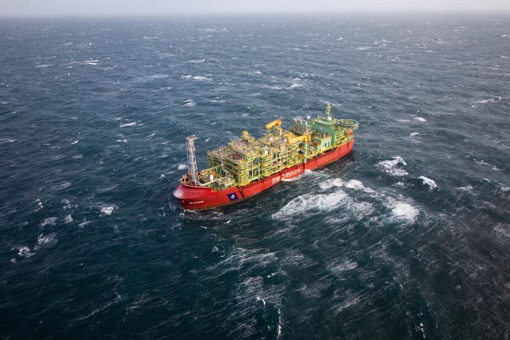 An aerial view of a large floating production storage and offloading (FPSO) vessel in turbulent ocean waters. The ship is predominantly red and yellow, equipped for processing and storing oil or gas.