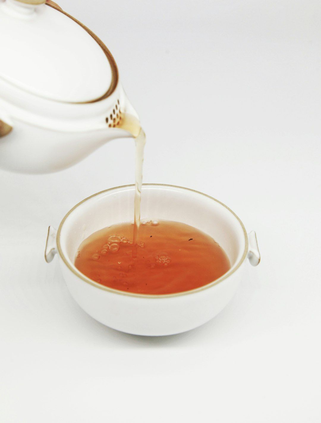 The image shows a teapot pouring tea into a white bowl. The tea is a warm amber color, suggesting it may be a traditional or herbal variety.