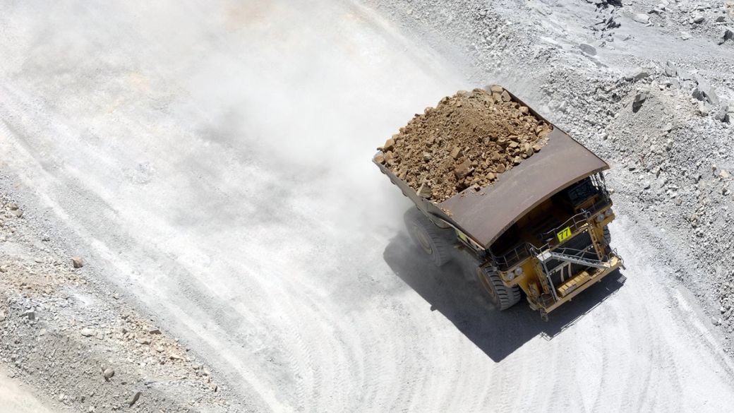 A large dump truck is seen transporting a load of rocks and gravel across a dusty construction site. The surroundings are dominated by pale, rocky terrain indicative of mining or excavation activities.