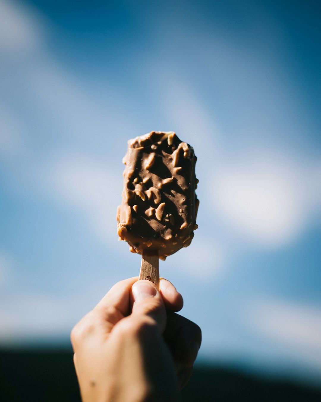 A hand holds a chocolate-covered ice cream bar on a stick against a bright blue sky. The ice cream is textured with nuts, suggesting a crunchy coating.
