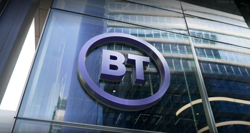 The image shows a close-up view of a modern office building's exterior featuring a prominent BT logo. Large glass windows reflect the surrounding environment, indicating a corporate setting.