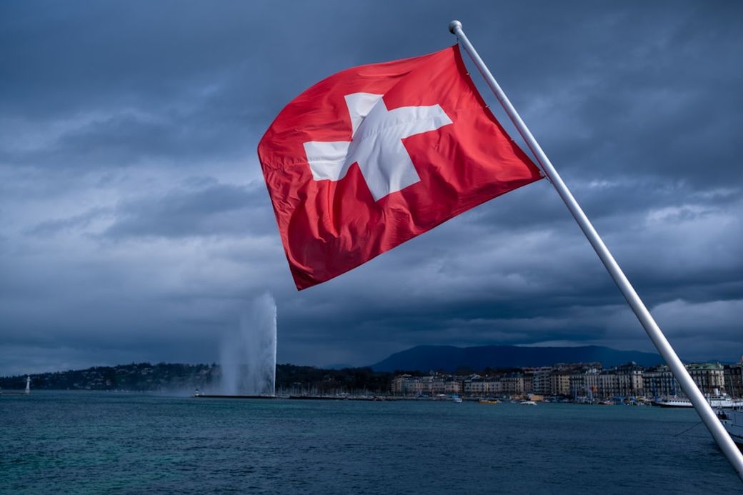 The image shows the Swiss flag prominently waving in the foreground, with a backdrop of a lake and a famous fountain in the distance. The sky is overcast, creating a dramatic atmosphere. — Credit: Photo by Ari Dinar on Unsplash