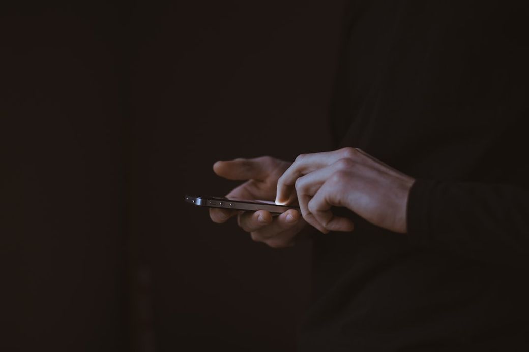 A person with partially visible hands is holding a smartphone in a dimly lit environment. The focus is on the smartphone, suggesting interaction or usage. — Credit: Photo by Gilles Lambert on Unsplash
