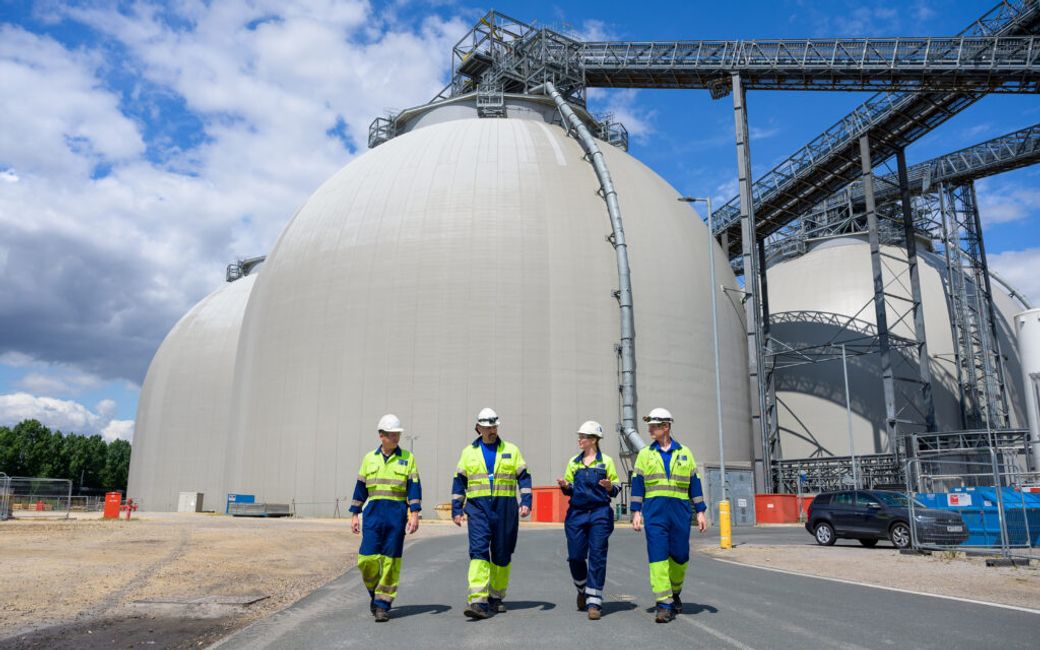 Four workers in safety gear walk together near large industrial storage tanks on a construction site. The tanks are likely part of an energy facility, with infrastructure visible in the background.