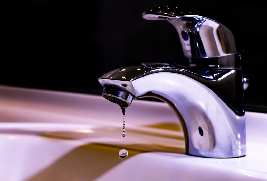 The image shows a modern chrome faucet with a water droplet hanging from the spout, set against a blurred dark background. The faucet is positioned over a white sink, emphasizing its sleek design. — Credit: Photo by Jos Speetjens on Unsplash