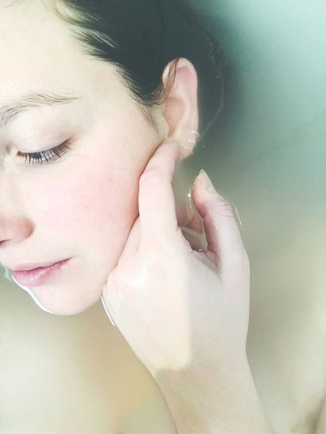 A close-up image of a woman partially submerged in water, with her face and hand visible as she touches her ear. The soft focus and natural lighting create a serene and calming atmosphere. — Credit: Photo by Coline Haslé on Unsplash