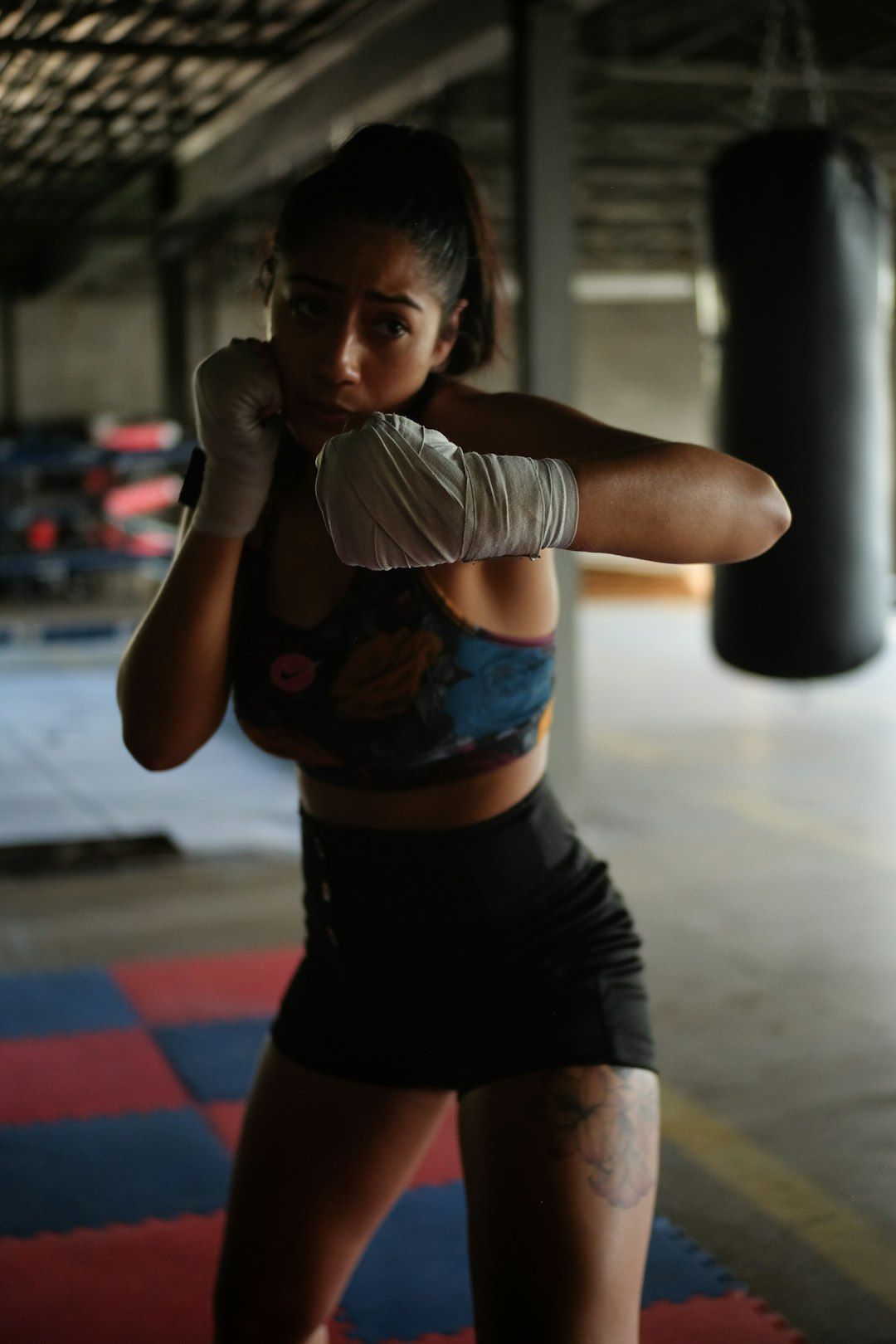 A female boxer is captured in a training gym, showcasing her readiness for a bout. She is in a fighting stance, with boxing gloves wrapped in tape and a determined expression.
