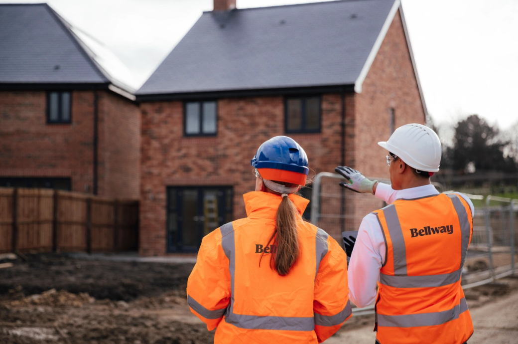 A construction site with two professionals observing a new residential building. One individual is wearing a hard hat and jacket branded with 'Bellway', while the other is in a white hard hat and reflective vest.