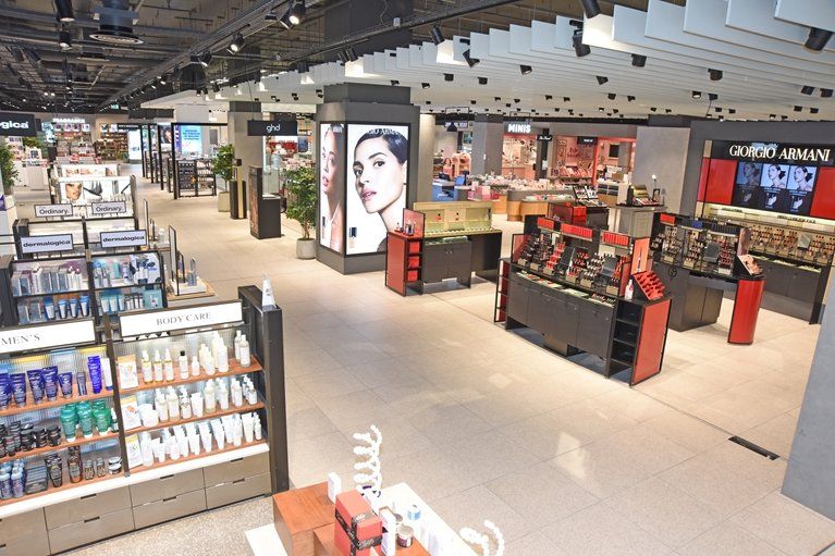 The image showcases a modern beauty retail store interior featuring various cosmetics and skincare products. Display stands are organized in a spacious layout with prominent brand signage visible on the walls and screens.
