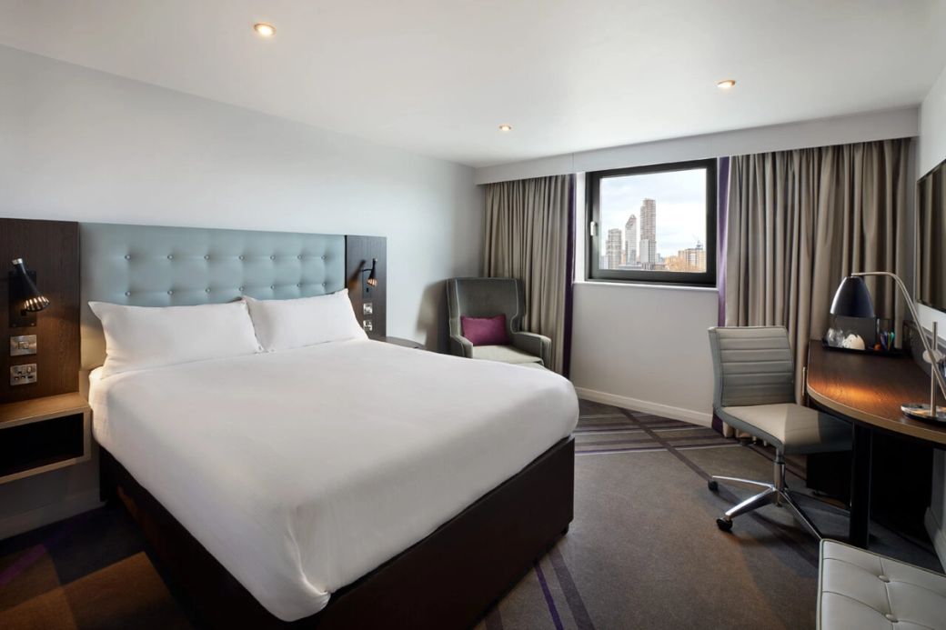 A modern hotel room featuring a neatly made king-sized bed, a stylish armchair by the window, and a workspace with a desk and chair. Natural light filters in through the window, highlighting the contemporary decor.