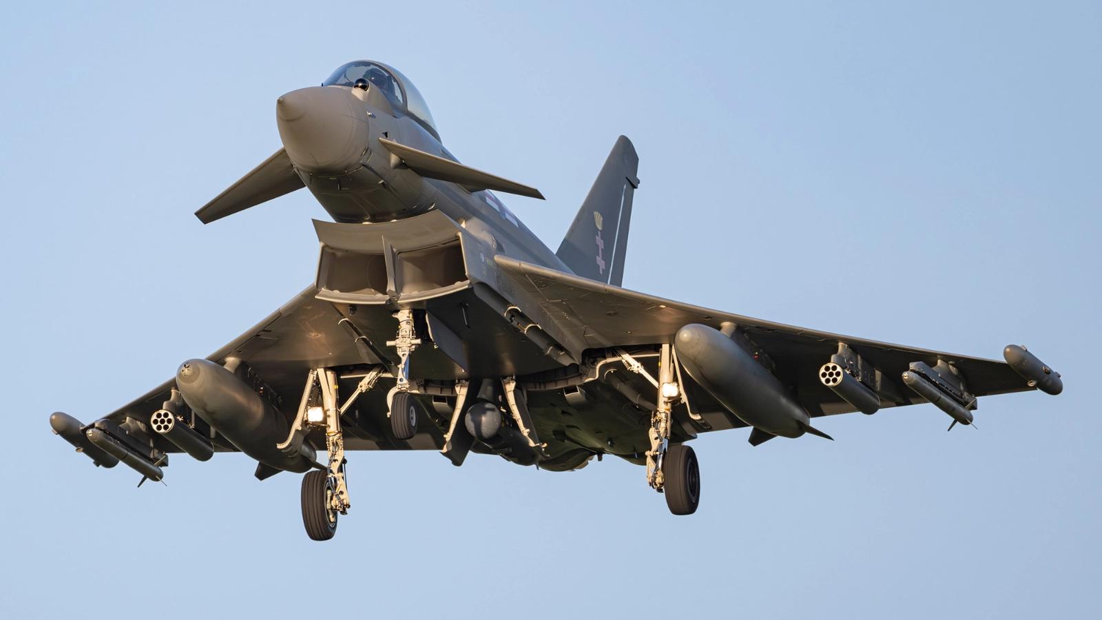 A military jet, specifically a Eurofighter Typhoon, is captured in flight against a clear blue sky. The aircraft is equipped with missiles and appears to be in a landing position.