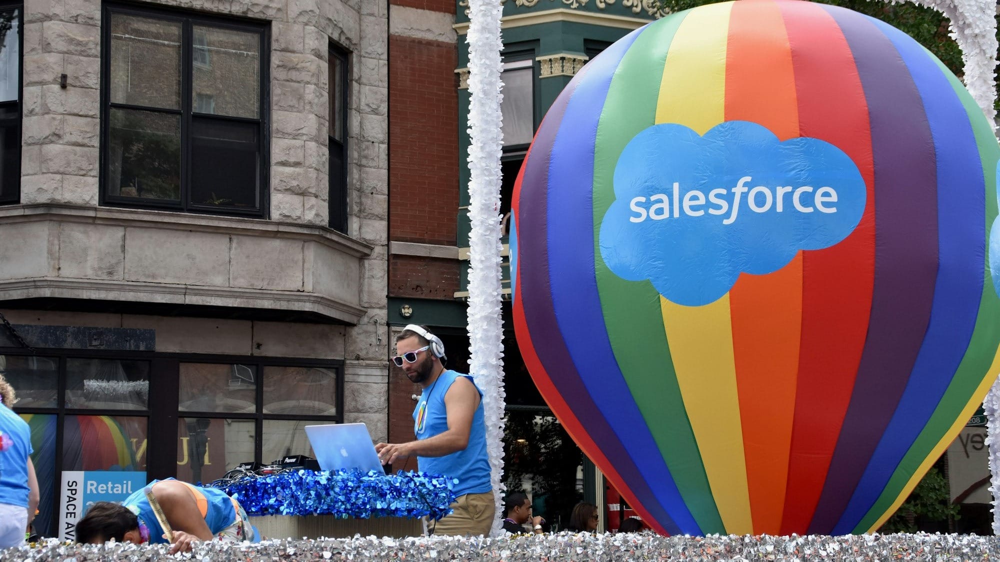 The image captures a vibrant parade scene featuring a DJ performing on a float, accompanied by a large colorful hot air balloon displaying the Salesforce logo. The atmosphere is festive and lively, indicative of a community celebration or event.
