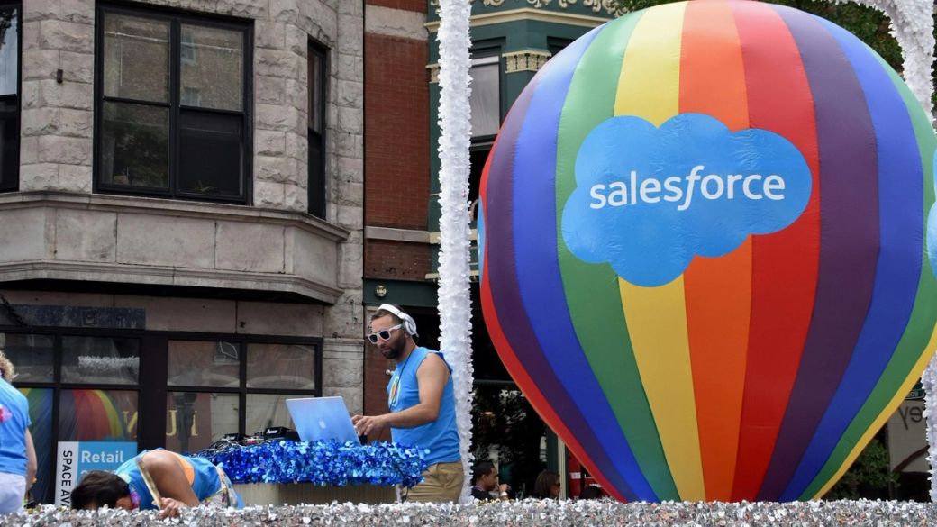 The image captures a vibrant parade scene featuring a DJ performing on a float, accompanied by a large colorful hot air balloon displaying the Salesforce logo. The atmosphere is festive and lively, indicative of a community celebration or event.