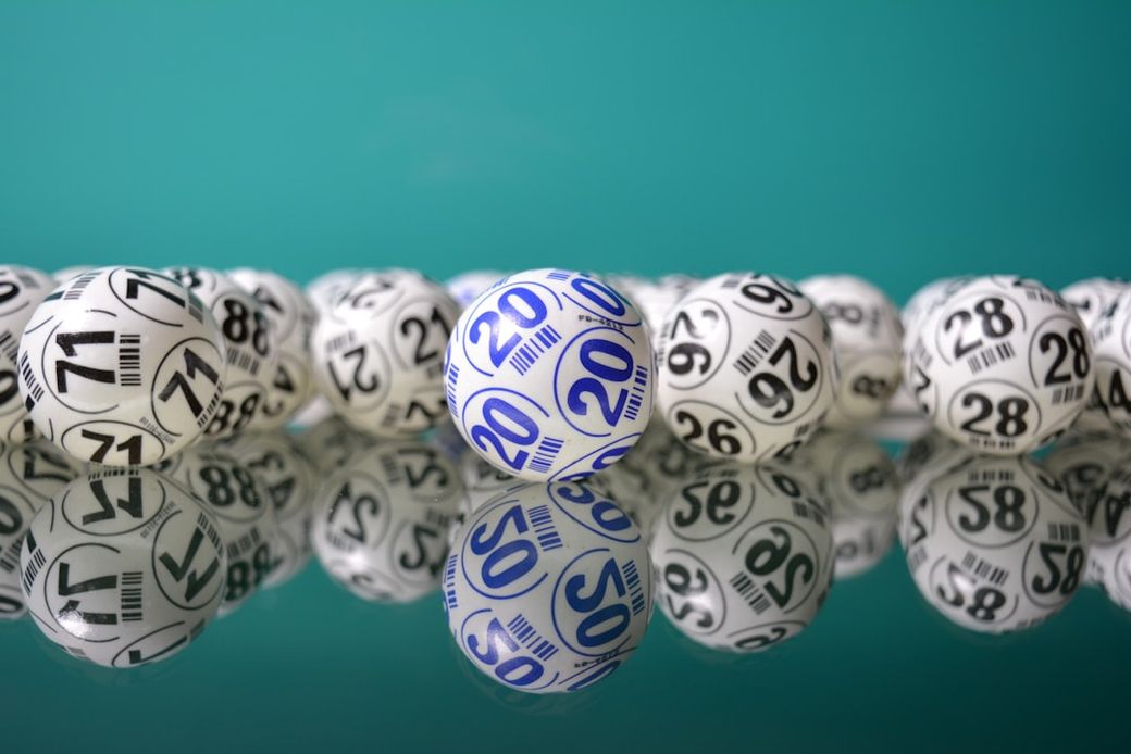 The image depicts a collection of numbered lottery balls arranged on a reflective surface. A blue ball marked with the number 20 stands out among a series of white balls displaying various numbers. — Credit: Photo by Alejandro Garay on Unsplash
