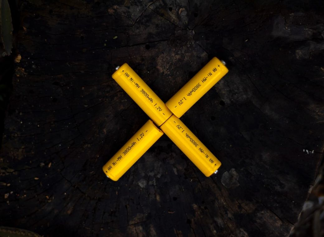The image features four yellow cylindrical batteries arranged in a cross formation on a dark wooden surface. The batteries are labeled with technical specifications, indicating their power capacity and type. — Credit: Photo by Saif71.com on Unsplash