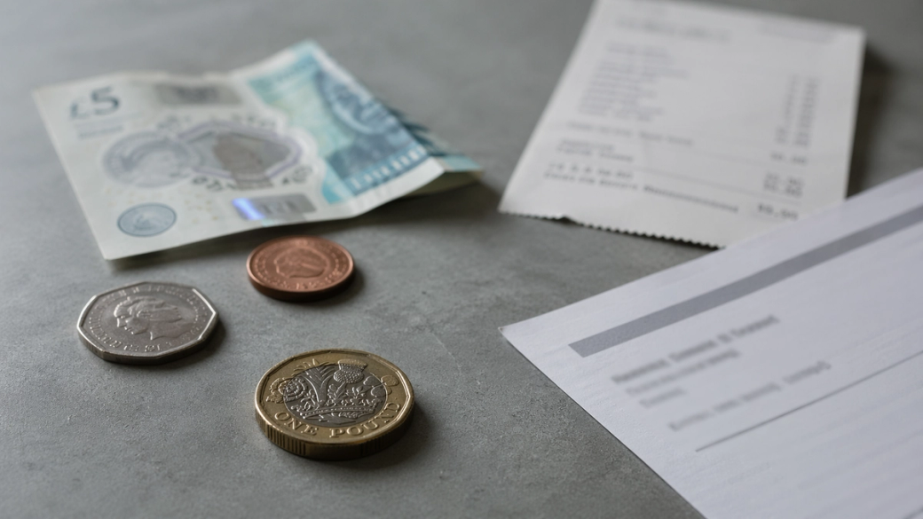 The image displays various denominations of British currency, including banknotes and coins, arranged on a concrete surface alongside a receipt. The visible notes include a five-pound note and a one-pound coin, highlighting aspects of currency and financial transactions.