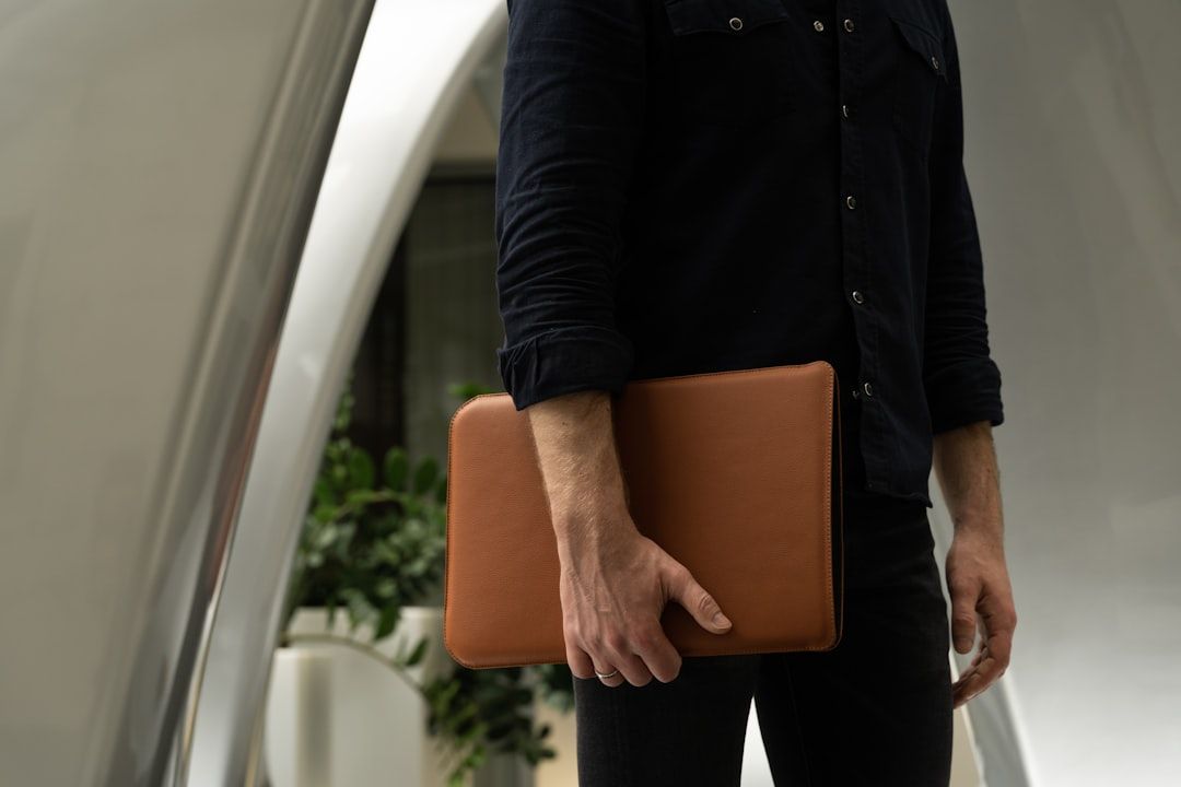 A man is standing in a modern architectural space, holding a brown leather laptop bag. The background features smooth, curved structures and greenery, suggesting a professional or corporate environment.