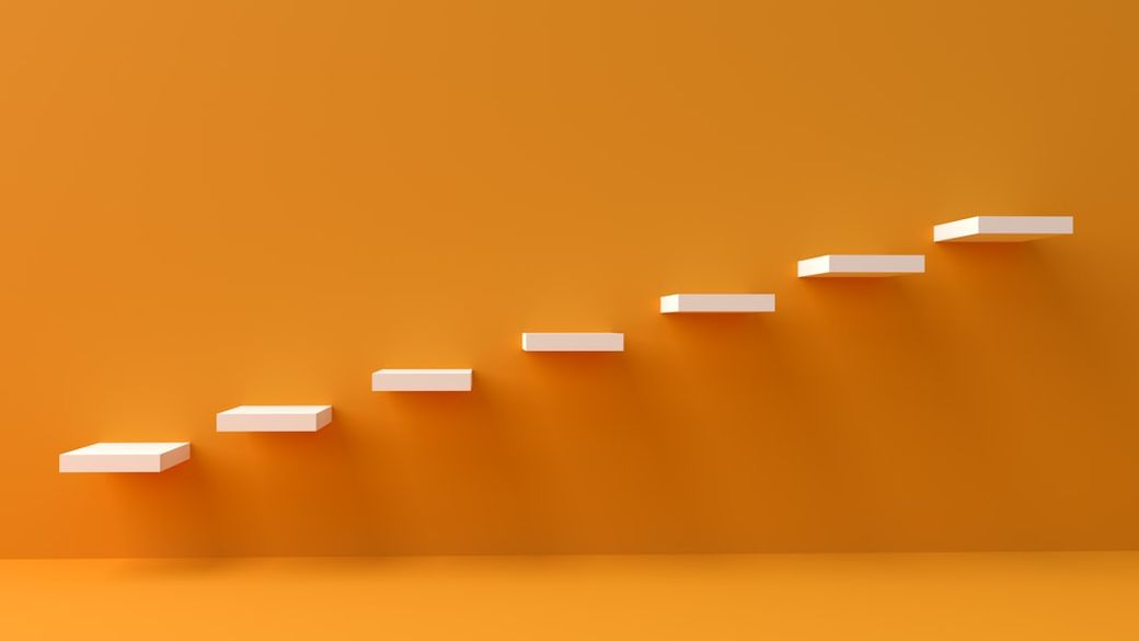 The image features a series of white, floating steps against a vibrant orange background. The steps ascend diagonally, creating a sense of progression or movement. — Credit: Photo by Kanhaiya Sharma on Unsplash