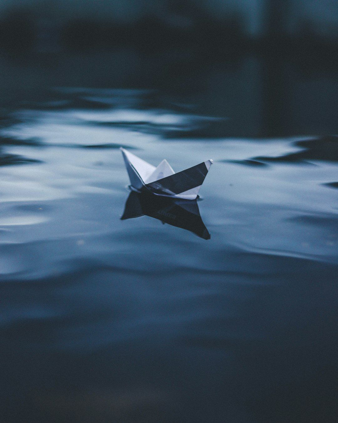 A small paper boat floats calmly on the surface of a dark, reflective body of water. The scene captures a serene moment with the paper boat slightly tilted, creating a peaceful ambiance.