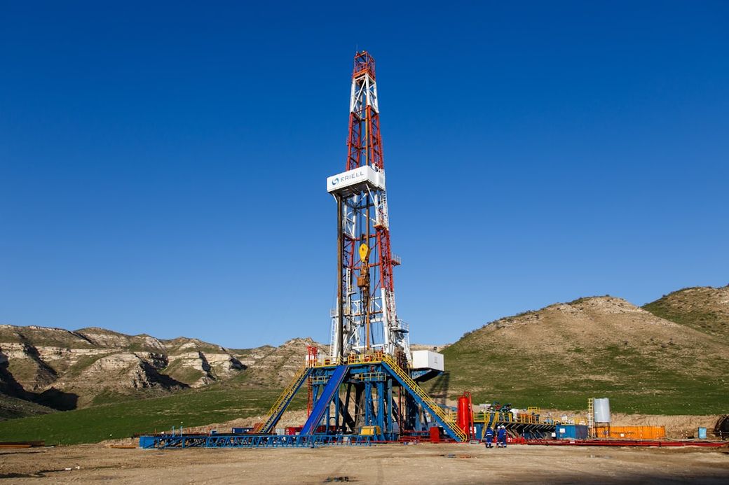 The image features a drilling rig set against a clear blue sky and rocky hills. The rig is prominently displayed in the foreground, showcasing its structure and equipment. — Credit: Photo by WORKSITE Ltd. on Unsplash