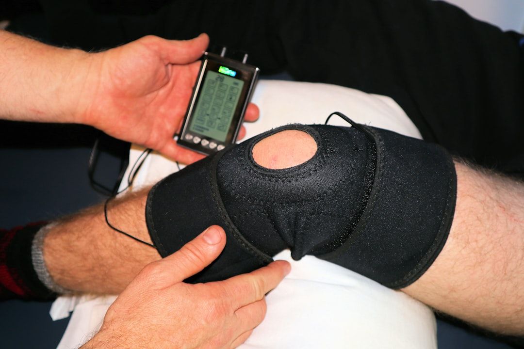 The image shows a close-up of a person's knee being examined with a diagnostic device. The knee is secured with a black support sleeve, and a hand is adjusting or monitoring the device.