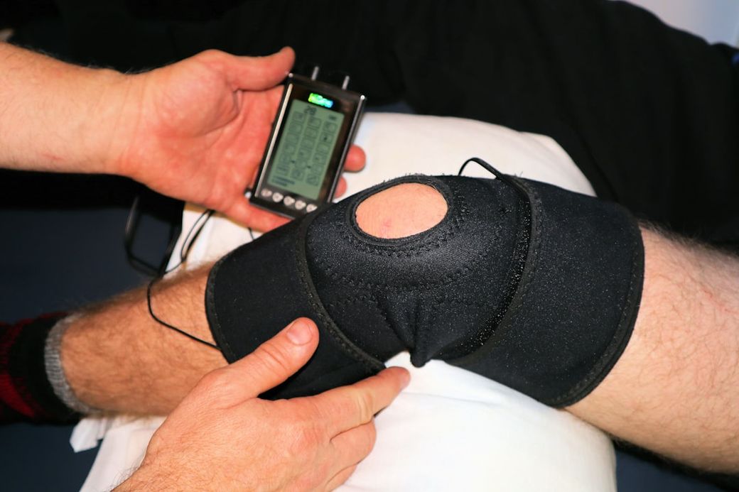 The image shows a close-up of a person's knee being examined with a diagnostic device. The knee is secured with a black support sleeve, and a hand is adjusting or monitoring the device. — Credit: Photo by Terry Shultz P.T. on Unsplash