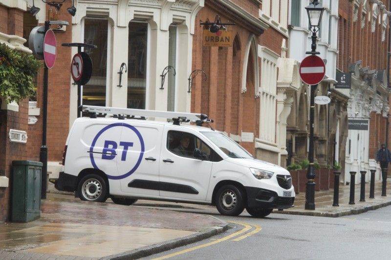 A white van with the brand 'BT' is parked at a street corner on a rainy day. The van is positioned near a building with brick architecture, surrounded by signage and street lamps.
