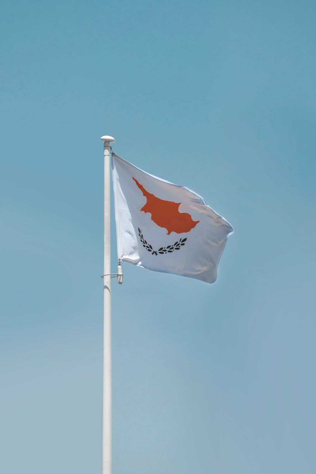 The image features the flag of Cyprus, prominently displayed on a flagpole. The flag showcases the map of Cyprus in orange, accompanied by olive branches below the map, against a clear blue sky.