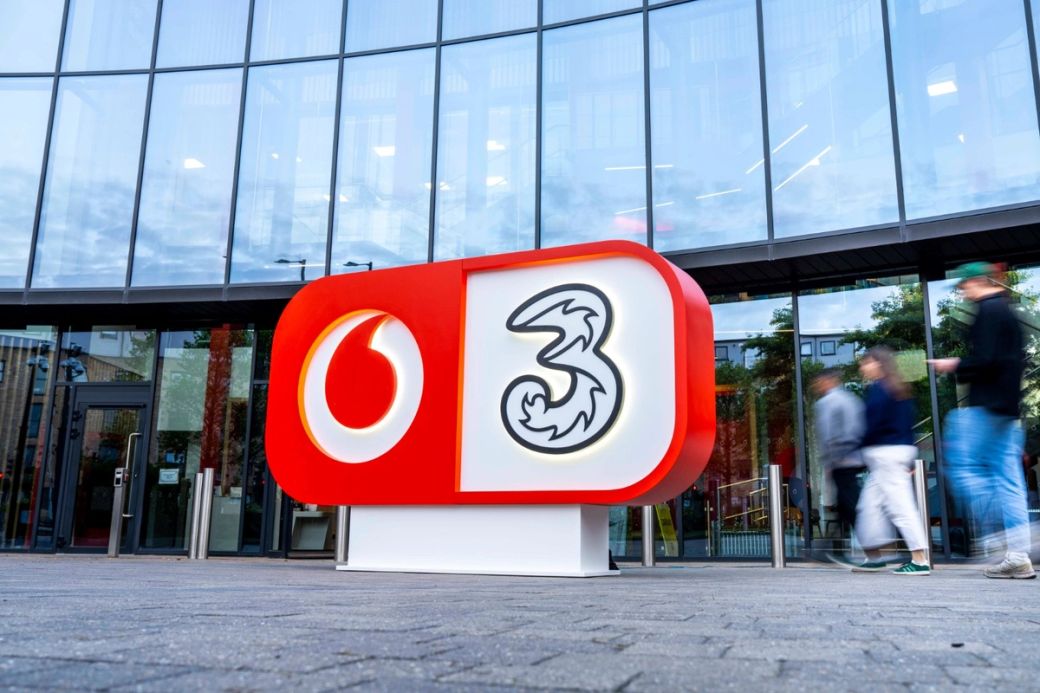 The image features a prominently displayed logo of Vodafone and 3 in front of a modern glass building. People can be seen walking by, indicating an active urban environment.
