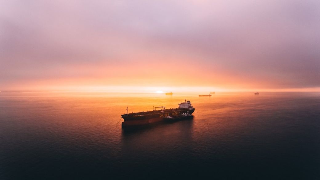 A large oil tanker is seen navigating through calm waters during sunrise, with multiple other vessels in the distance. The sky is filled with soft hues of pink and orange as the sun rises above the horizon. — Credit: Photo by Ian Simmonds on Unsplash