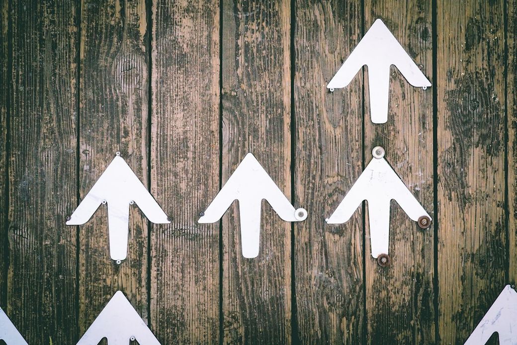 The image features several white arrow signs arranged on a wooden surface. The arrows point in various directions, suggesting movement or guidance, and they are placed against a textured brown backdrop. — Credit: Photo by Jungwoo Hong on Unsplash