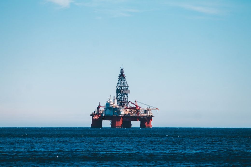 An offshore oil drilling rig stands prominently in the open sea against a clear blue sky. The structure is equipped with various machinery, indicative of its purpose in oil extraction and energy production. — Credit: Photo by Erik Mclean on Unsplash