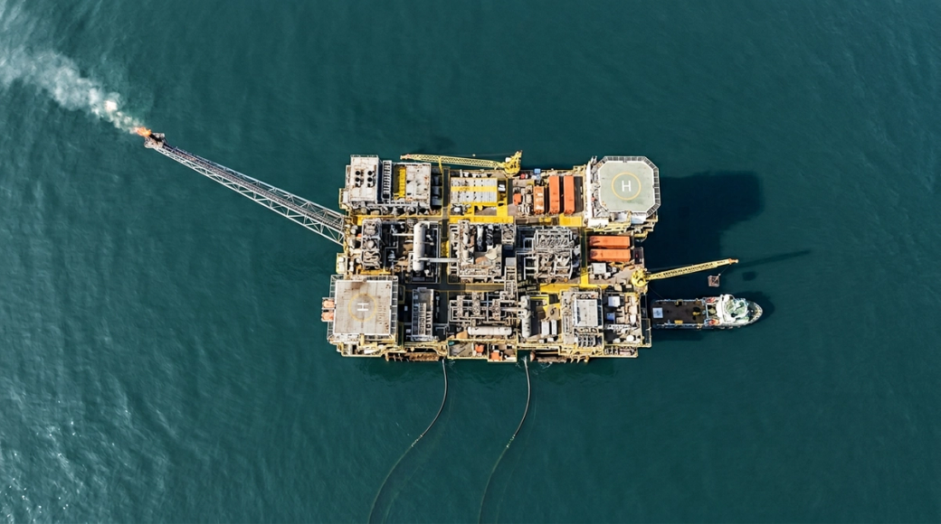 The image shows a high aerial view of a fixed production platform complex situated on calm teal waters. The intricate layout includes pipe racks, separator vessels, and accommodation blocks arranged in functionally distinct zones, highlighted by a mix of weathered grey rooftops, safety yellow walkways, and orange container units. A supply vessel is moored alongside, with a crane arm positioned above it, while the flare boom extends diagonally, creating a striking industrial composition.