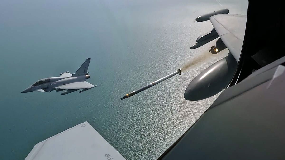 The image depicts a military aircraft firing a missile over a body of water, showcasing a dynamic aerial view. The jet is positioned in the foreground while the missile is mid-launch, highlighting the action of modern military operations.