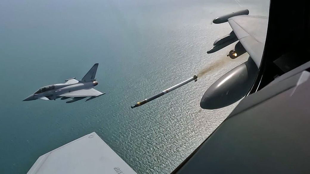 The image depicts a military aircraft firing a missile over a body of water, showcasing a dynamic aerial view. The jet is positioned in the foreground while the missile is mid-launch, highlighting the action of modern military operations.