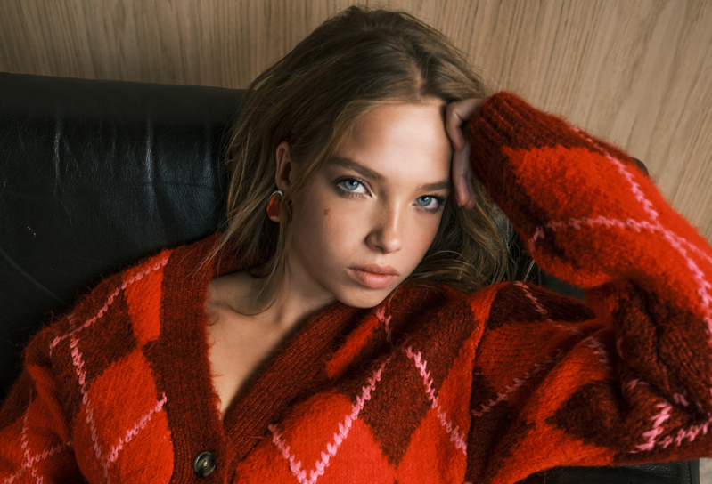 A woman is posing on a black leather couch, wearing a stylish red and pink patterned sweater. She gazes intently at the camera, with her hand resting on her forehead.
