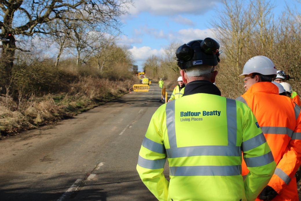 The image depicts a group of construction workers and officials gathered on a road where roadworks are taking place. One worker in the foreground is wearing a jacket with 'Balfour Beatty Living Places' printed on it, indicating their affiliation.