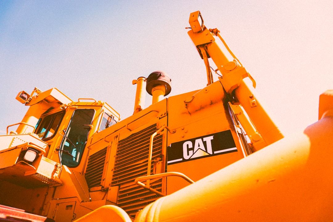 The image features a close-up view of a yellow construction vehicle, prominently showcasing its machinery and structure against a clear blue sky. The foreground highlights details of the vehicle's design, including its operator cabin and arm.