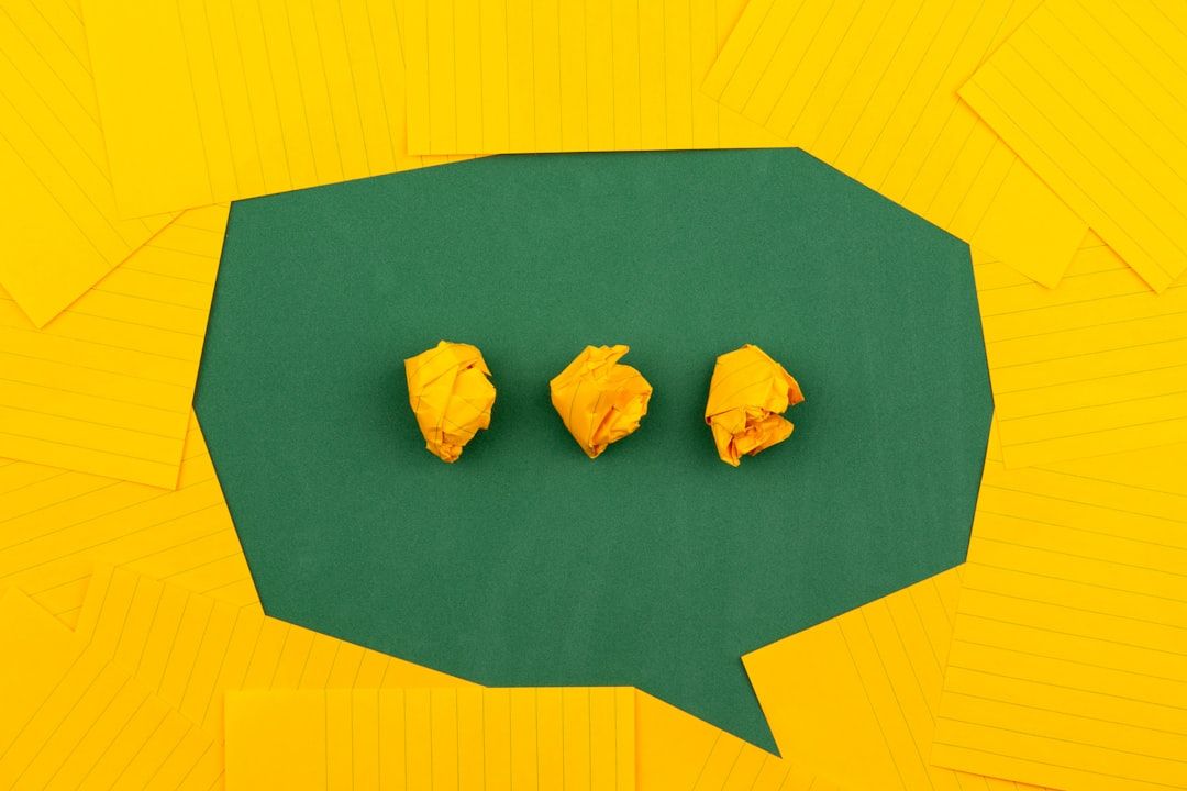 The image features three crumpled yellow paper balls arranged on a green surface that is shaped like a speech bubble. Surrounding the bubble are scattered sheets of lined yellow paper, creating a vibrant and creative atmosphere.