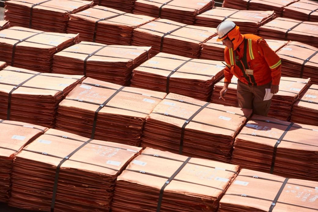 The image shows a worker in safety gear inspecting large bundles of copper sheets stacked neatly in a warehouse or storage area. The bright orange color of the copper contrasts with the worker's orange safety vest and helmet.